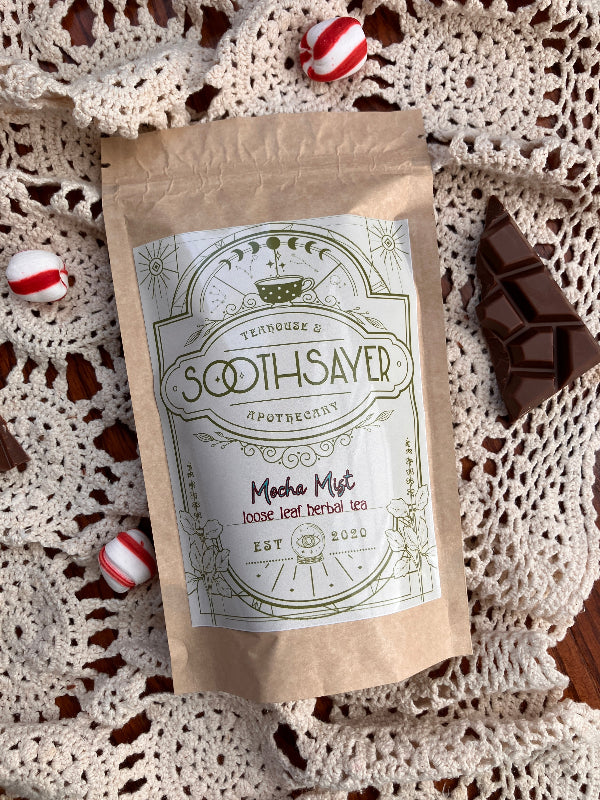 Tea package labeled 'Soothsayer' on a crocheted surface with candies and chocolate.