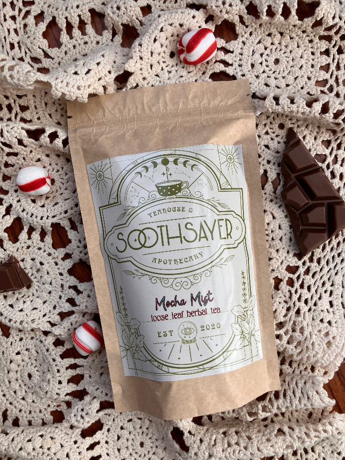 Tea package labeled 'Soothsayer' on a lace doily with chocolate and peppermint candy.
